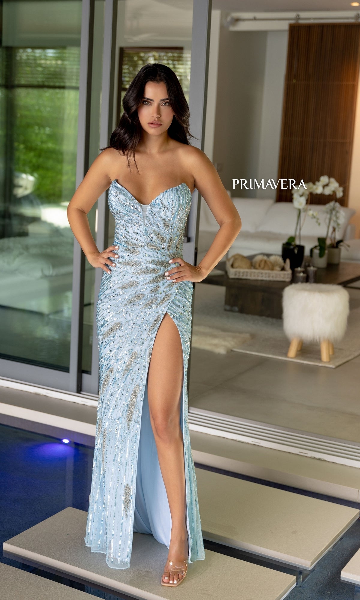 A woman wears the Strapless Primavera Long Beaded Formal Dress 4108 with a thigh-high slit, standing indoors on a platform, hands on hips.